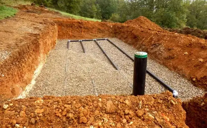 An open drain field trench cut into red clay soil, with perforated pipes laid on a gravel bed